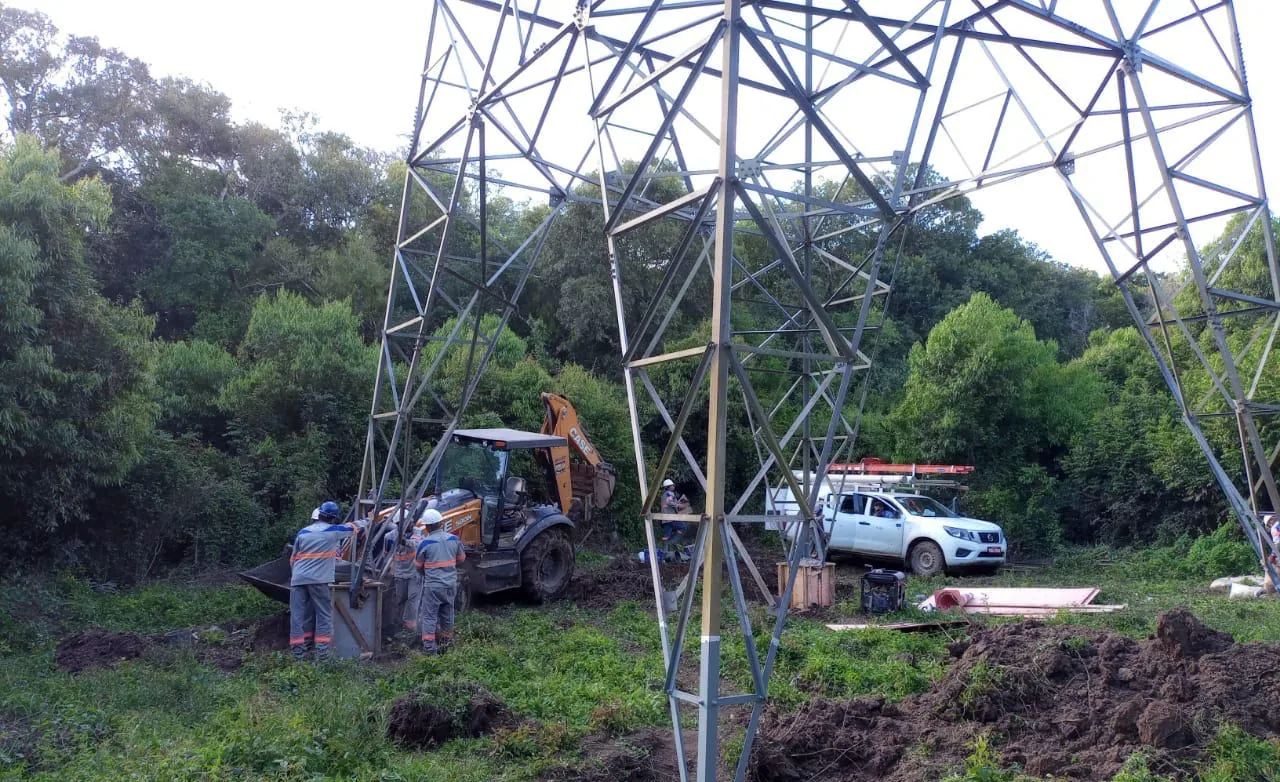 Equipe e retroescavadeira trabalhando na base de uma torre de transmissão em área de mata