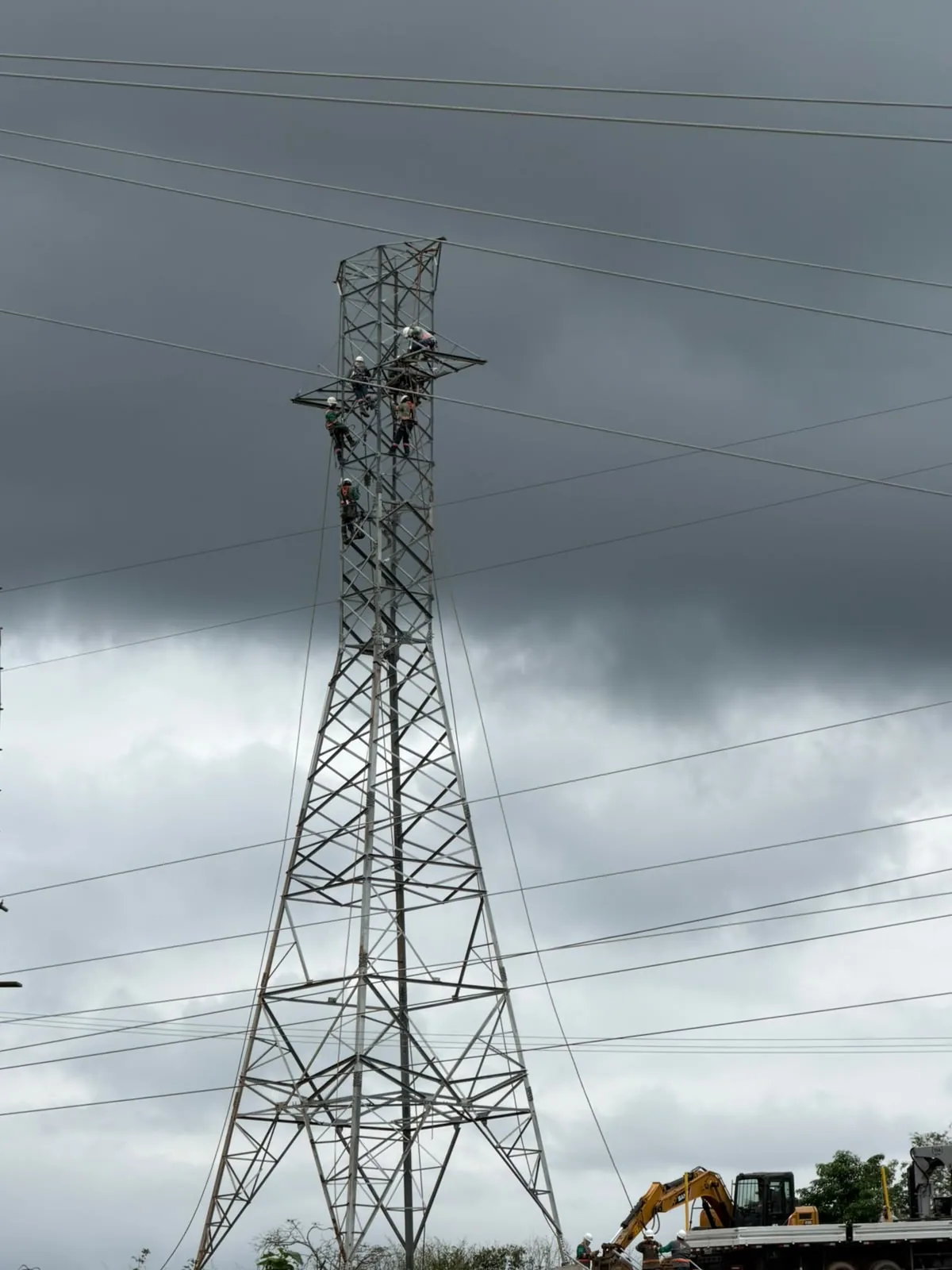 Torre de transmissão com equipe no alto e escavadeira na base